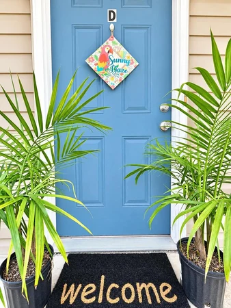 Blue front door marked D, with a welcome mat and potted palms